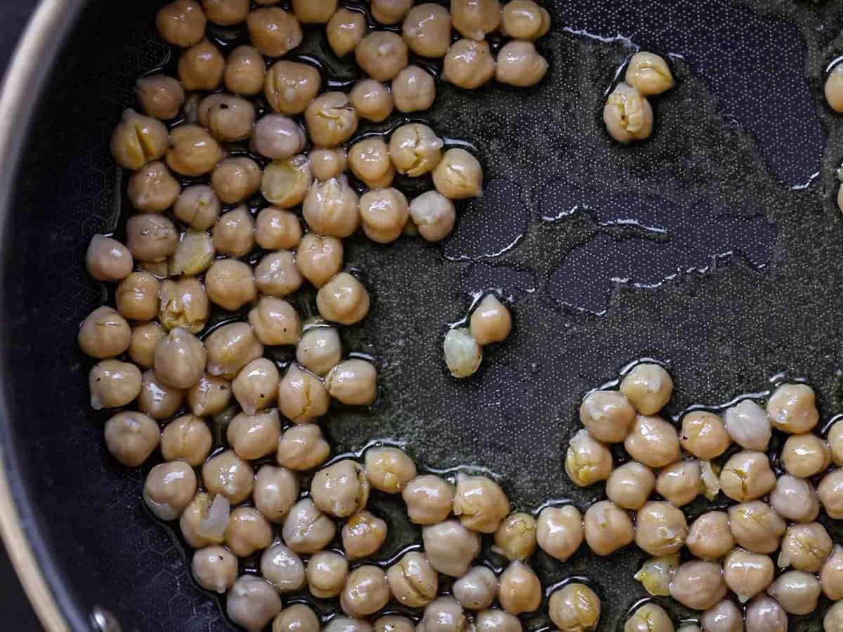 Chickpeas being sautéed in a black frying pan with a small amount of oil. The chickpeas are spread out, and the pans surface is visible.